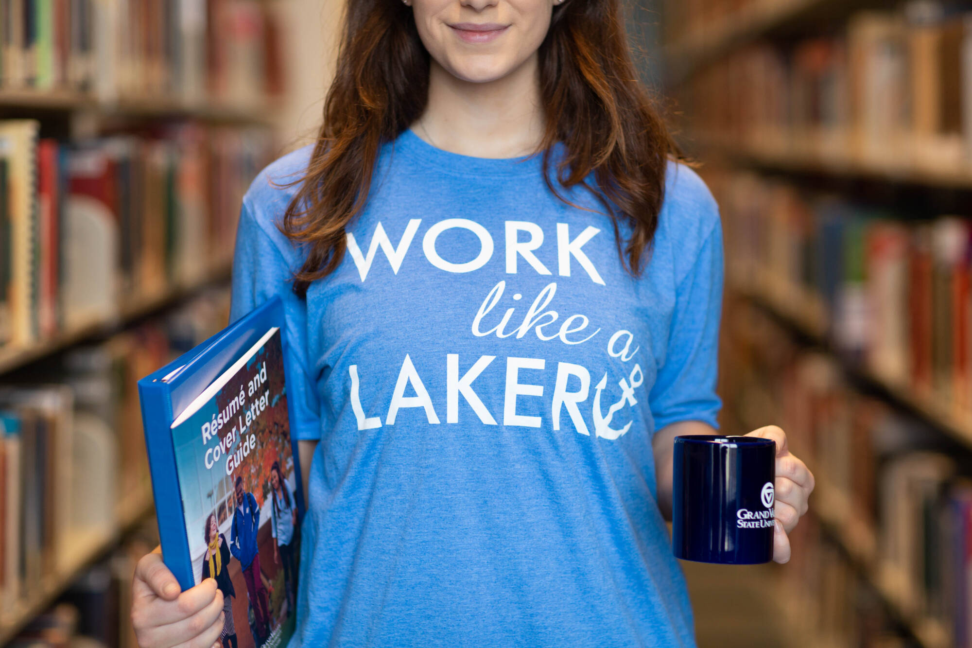 A young woman in a blue t-shirt that says "WORK like a LAKER" holds a binder and a mug in a library setting.